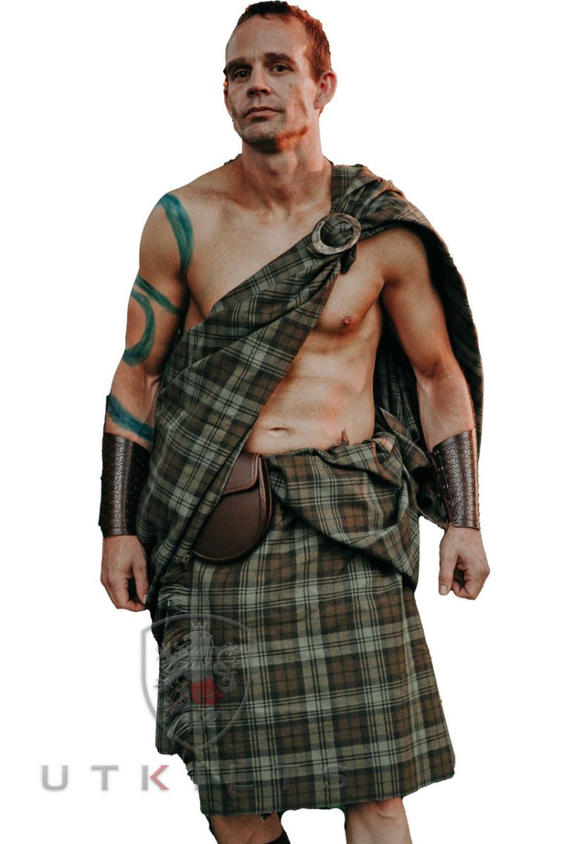 A man in a kilt stands confidently against a white background, showcasing traditional Scottish attire.